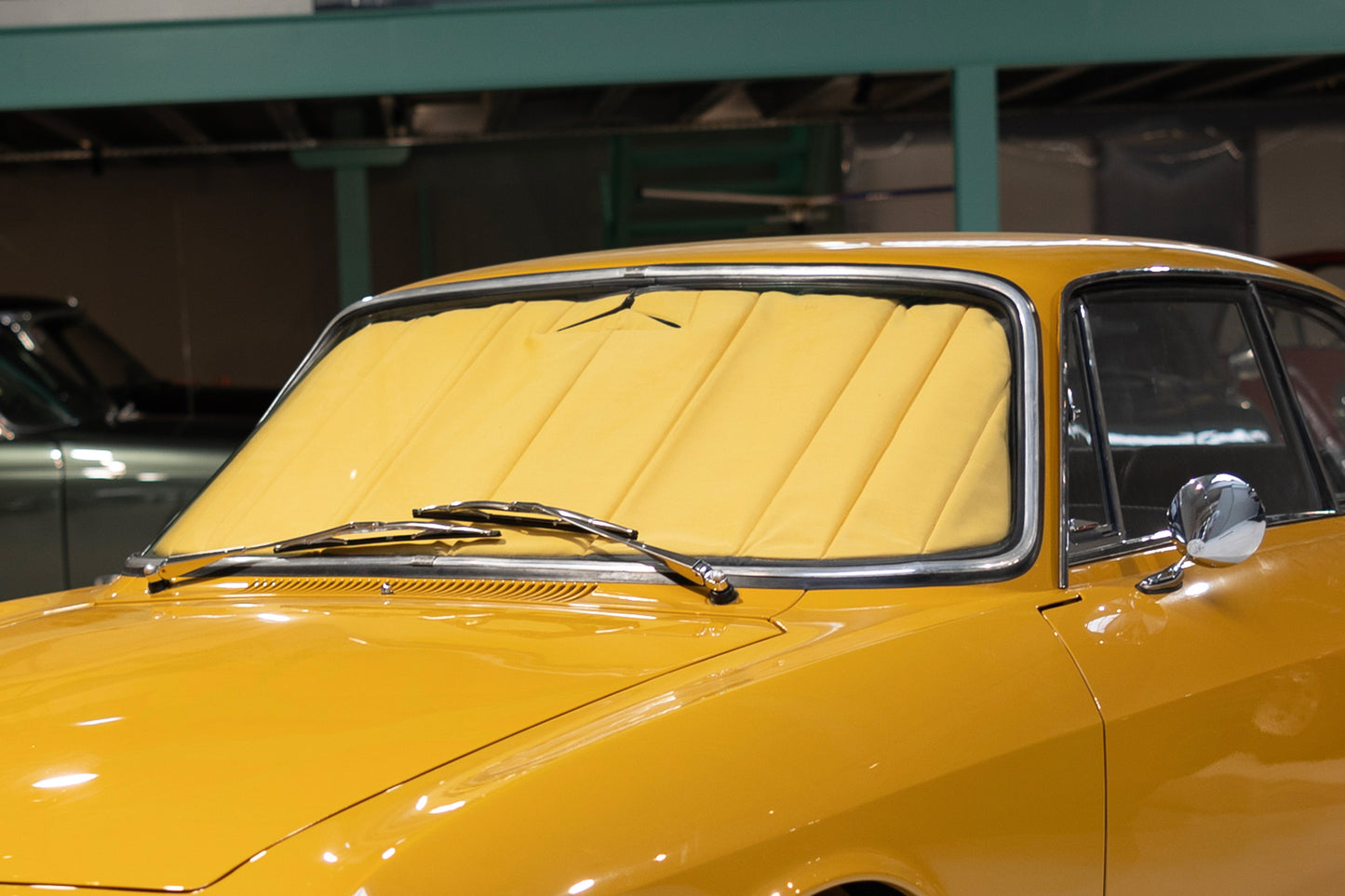 Classic yellow car fitted with a standard premium beige sunshade, emphasizing interior protection and timeless design in a showroom setting. Autentici sun shade for car windshield. Protetor solar para para-brisas de automóvel clássico.