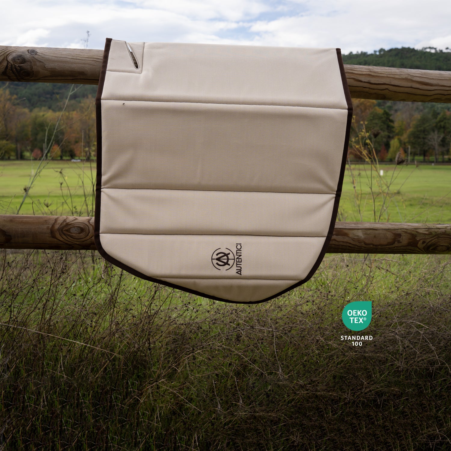 Beige premium car sunshade with brown trim and embroidered logo displayed on a wooden fence in a natural setting, certified OEKO-TEX Standard 100 for eco-friendly quality and sustainability. Autentici sun shade for car windshield. Protetor solar para para-brisas de automóvel clássico.