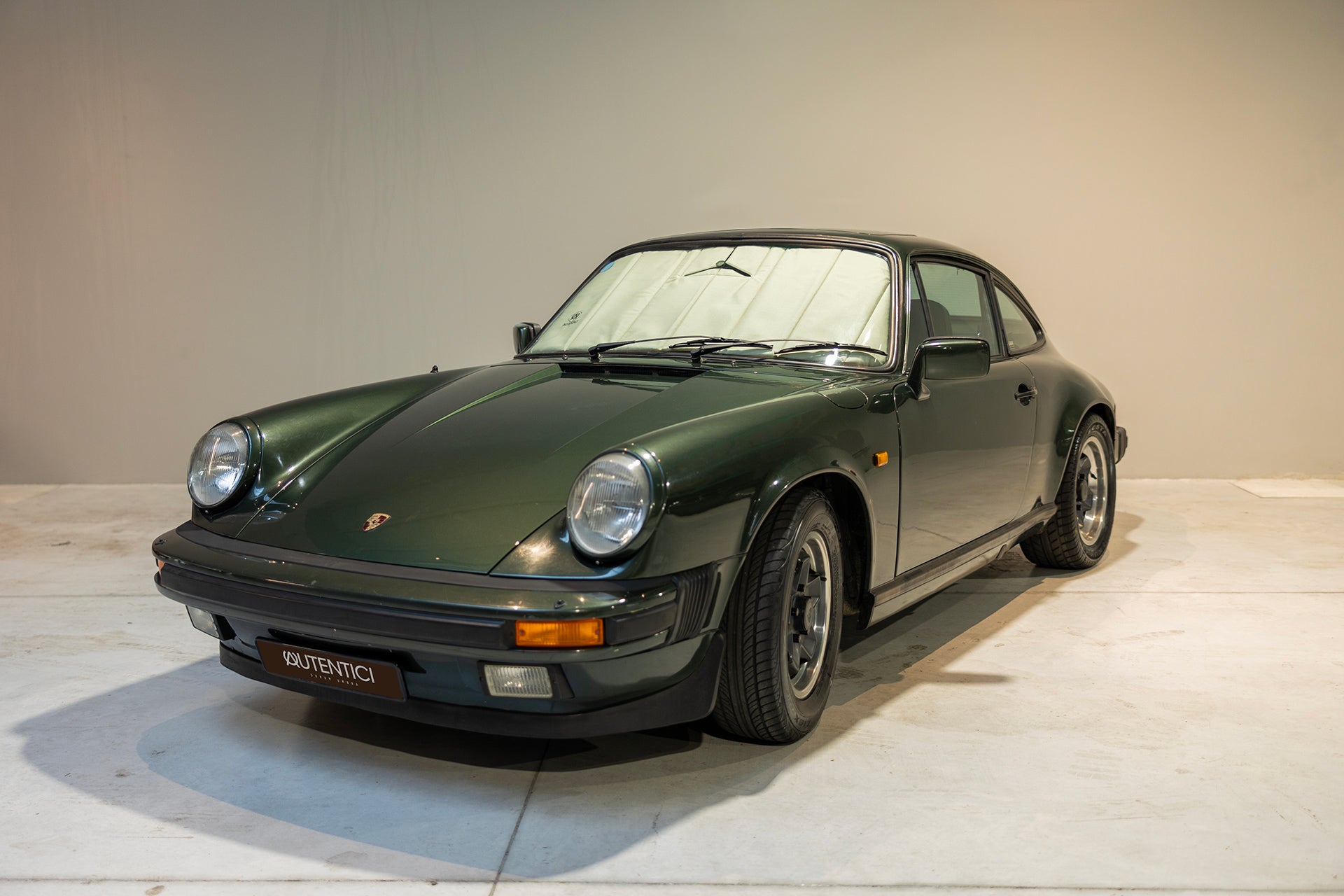 Classic green car fitted with a premium beige sunshade, emphasizing tailored interior protection and timeless elegance in a showroom setting.  Autentici sun shade for car windshield. Protetor solar para para-brisas de automóvel clássico.