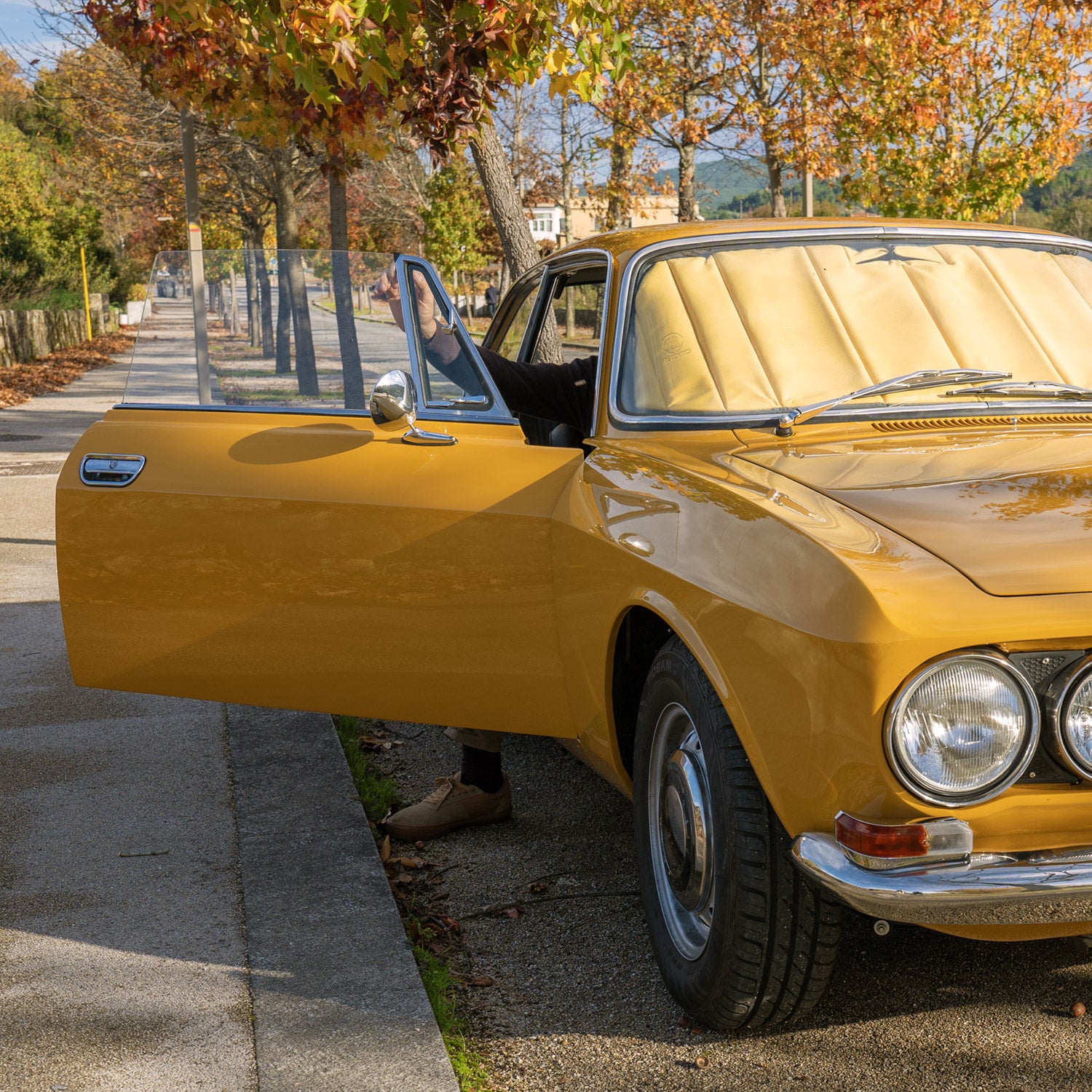 Classic yellow car with an open door, featuring a premium sunshade on the windshield, parked on an autumn tree-lined street, showcasing timeless elegance and custom car interior protection, Autentici sun shade for car windshield. Protetor solar para para-brisas de automóvel clássico.