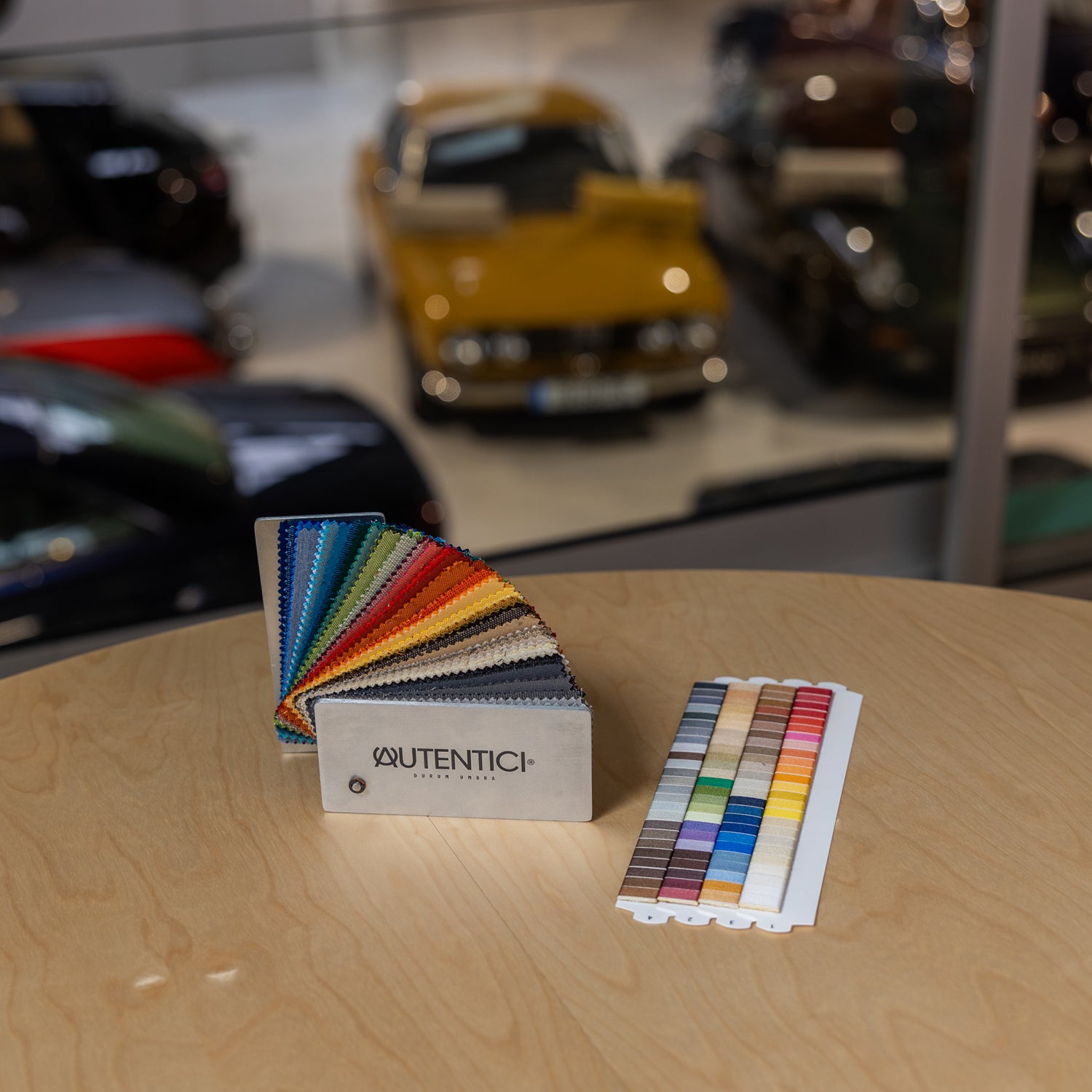 Fabric color samples displayed on a wooden table with classic cars in the background, showcasing customization options for premium car sunshades and tailored interior accessories. Autentici sun shade for car windshield. Protetor solar para para-brisas de automóvel clássico.