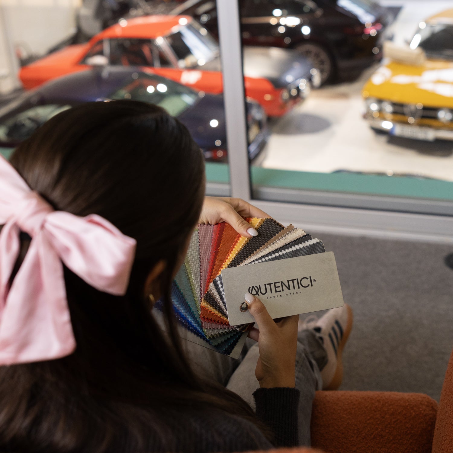  Person holding a fabric color sample booklet with customization options for premium car sunshades, overlooking a showroom with classic cars in the background. Autentici sun shade for car windshield. Protetor solar para para-brisas de automóvel clássico.