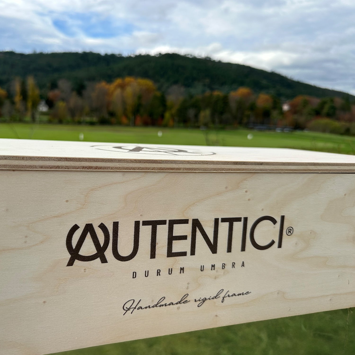 Wooden packaging with an engraved logo for a handmade rigid frame car accessory, set against a scenic countryside background, showcasing premium craftsmanship and sustainability. Autentici sun shade for car windshield.